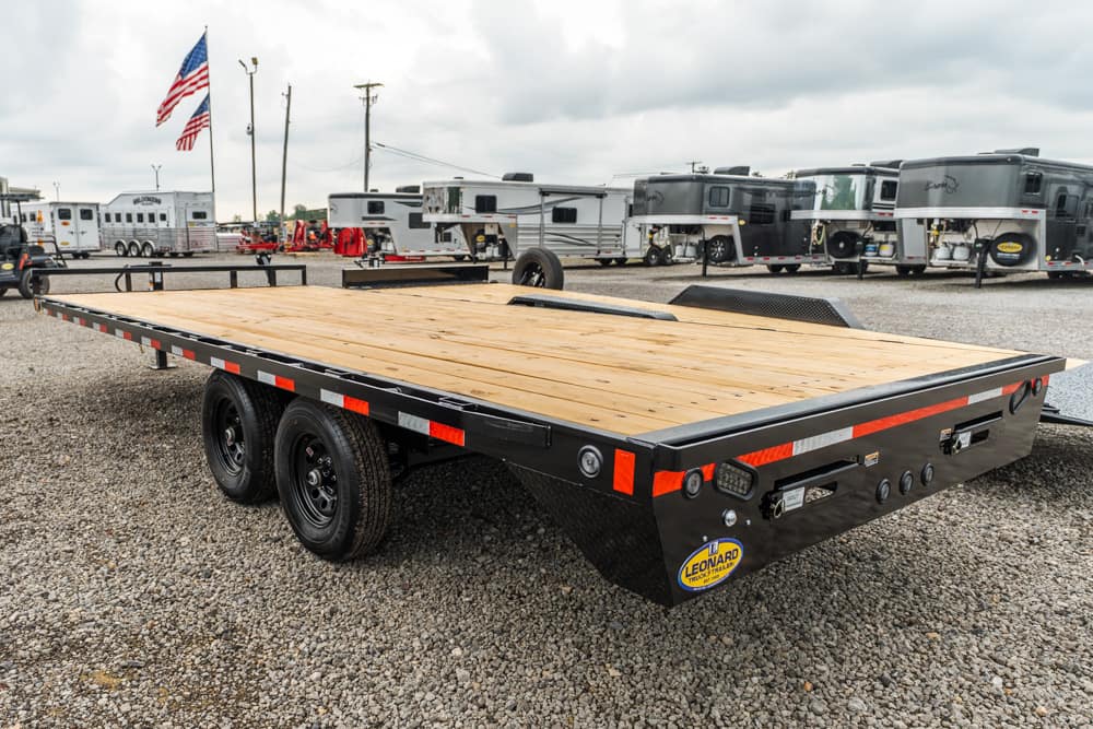 Dual-axle flatbed trailer with a wooden deck in a gravel RV lot, with an American flag and several campers in the background.