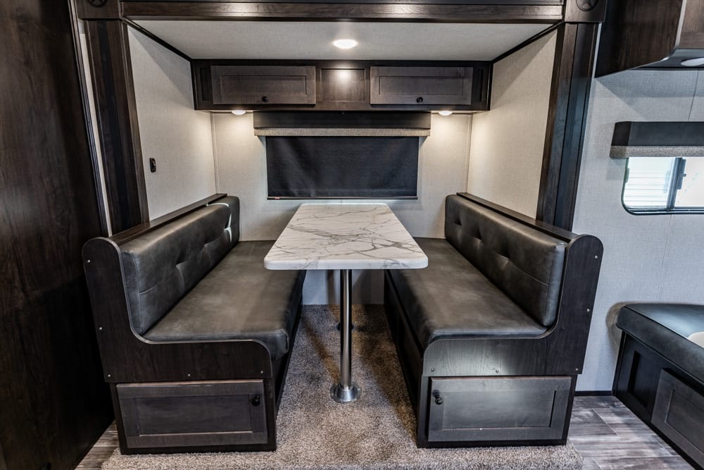 RV dinette booth with dark leatherette benches and a marble-patterned table, window shade behind, overhead cabinets above