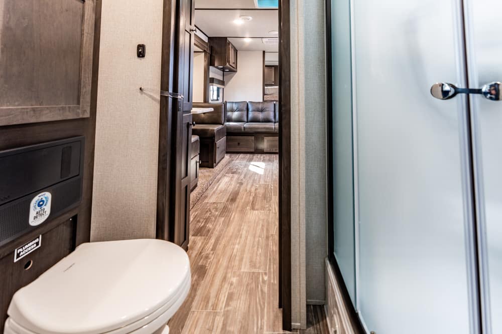 Interior of an RV/trailer hallway: toilet on the left, doorway to a living area with a black leather sofa ahead, glass shower on the right edge, wood-like flooring throughout