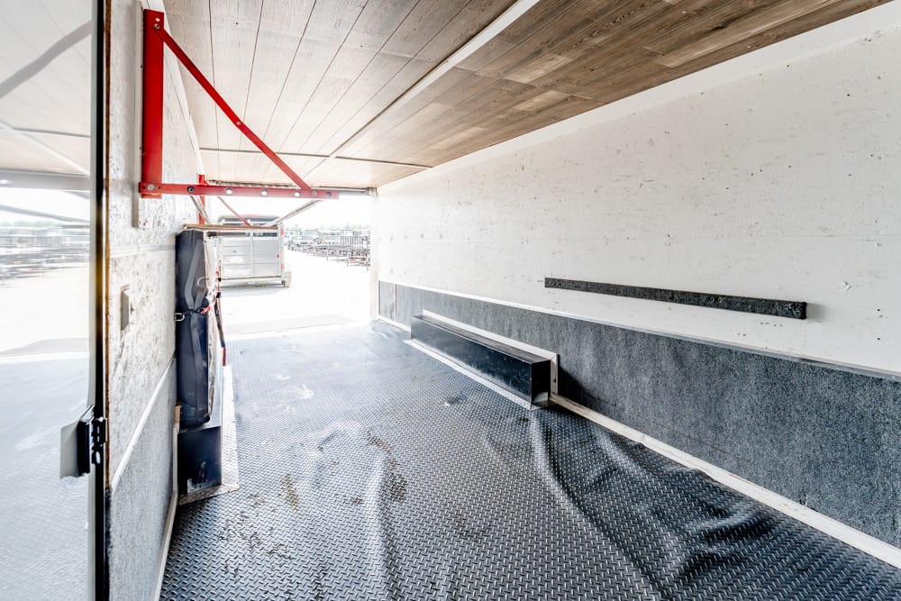 Interior of an empty cargo trailer with a red wall brace, textured metal floor, and open rear doors showing a loading area outside.