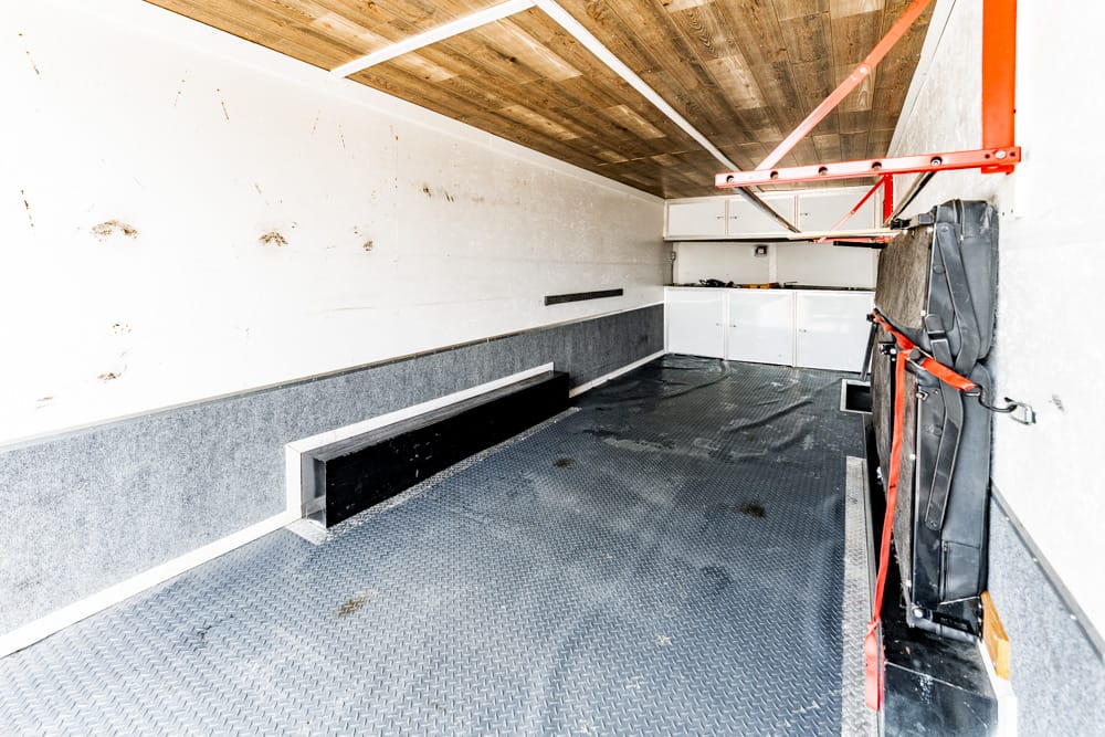 Interior of an empty utility trailer with diamond-plate floor, white walls, and cabinets at the far end.