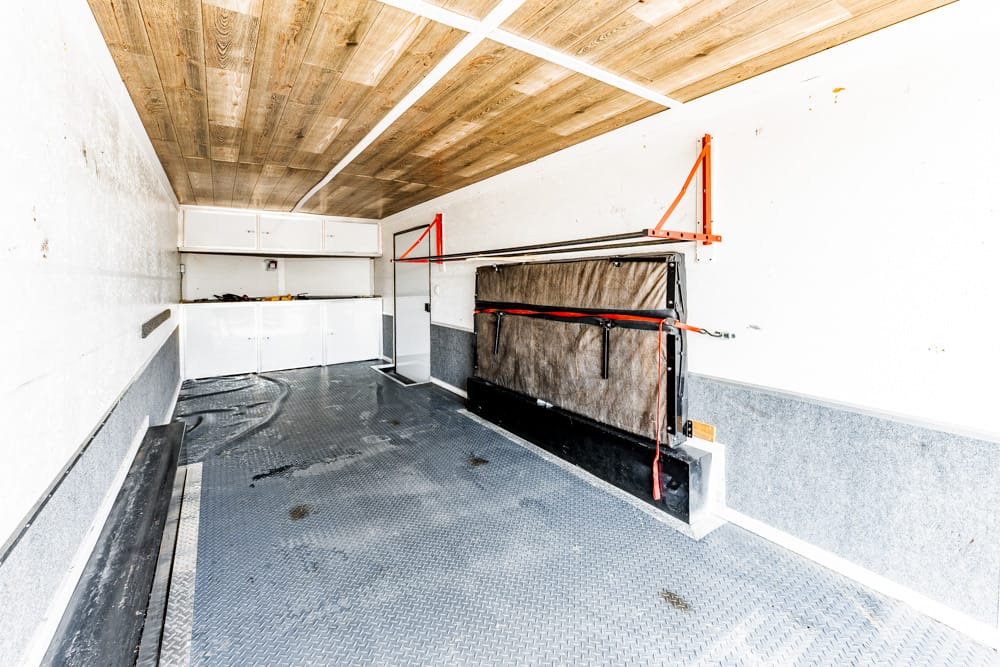 Interior of an empty utility trailer with a folded, strapped-down mattress on the right, diamond-plate floor, white walls, and a wooden ceiling.