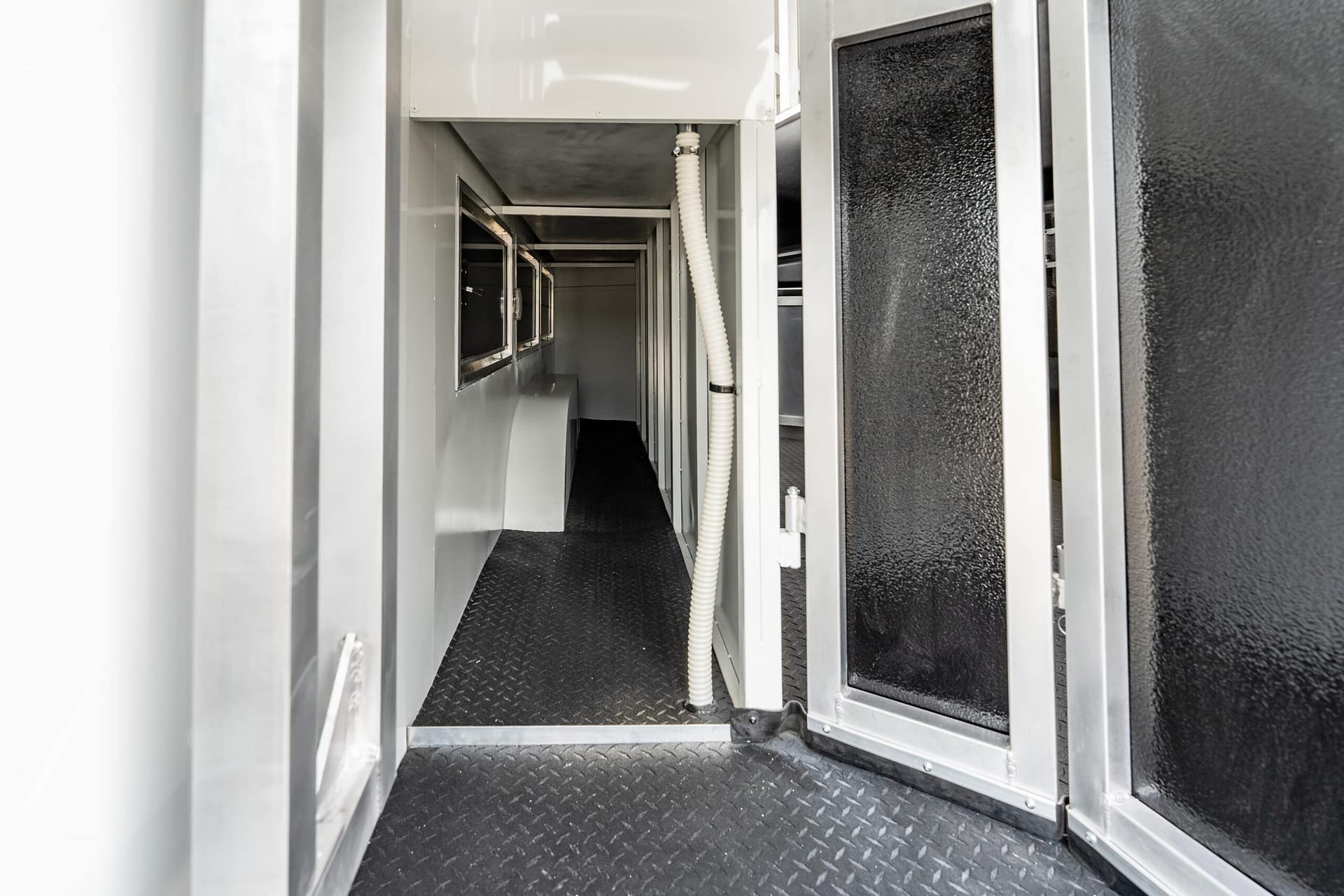 Narrow industrial corridor with metal framing, a white corrugated hose running along the wall, and a diamond-plate floor.