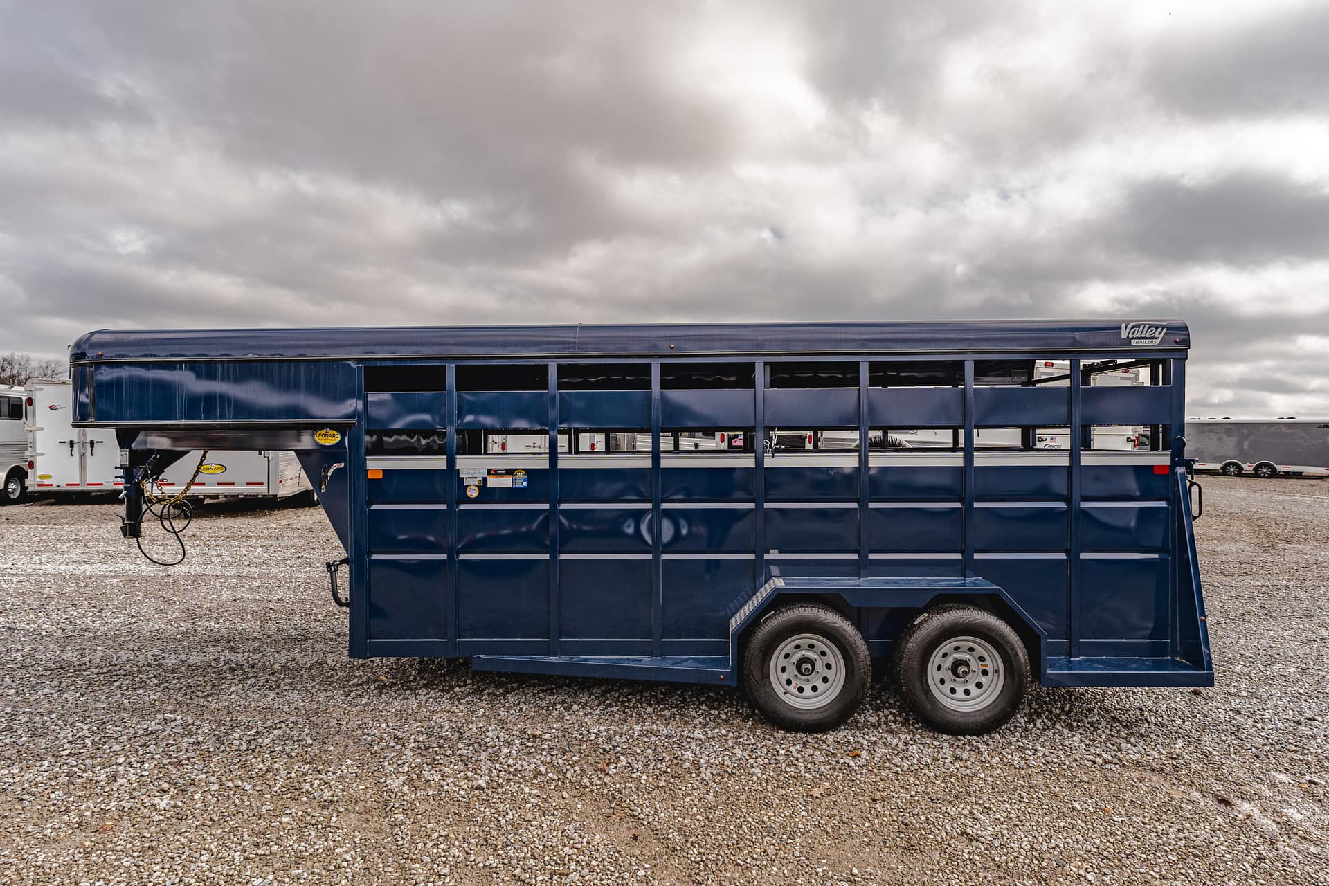 Blue long livestock trailer on a gravel lot under a cloudy sky, with other trailers visible in the background.