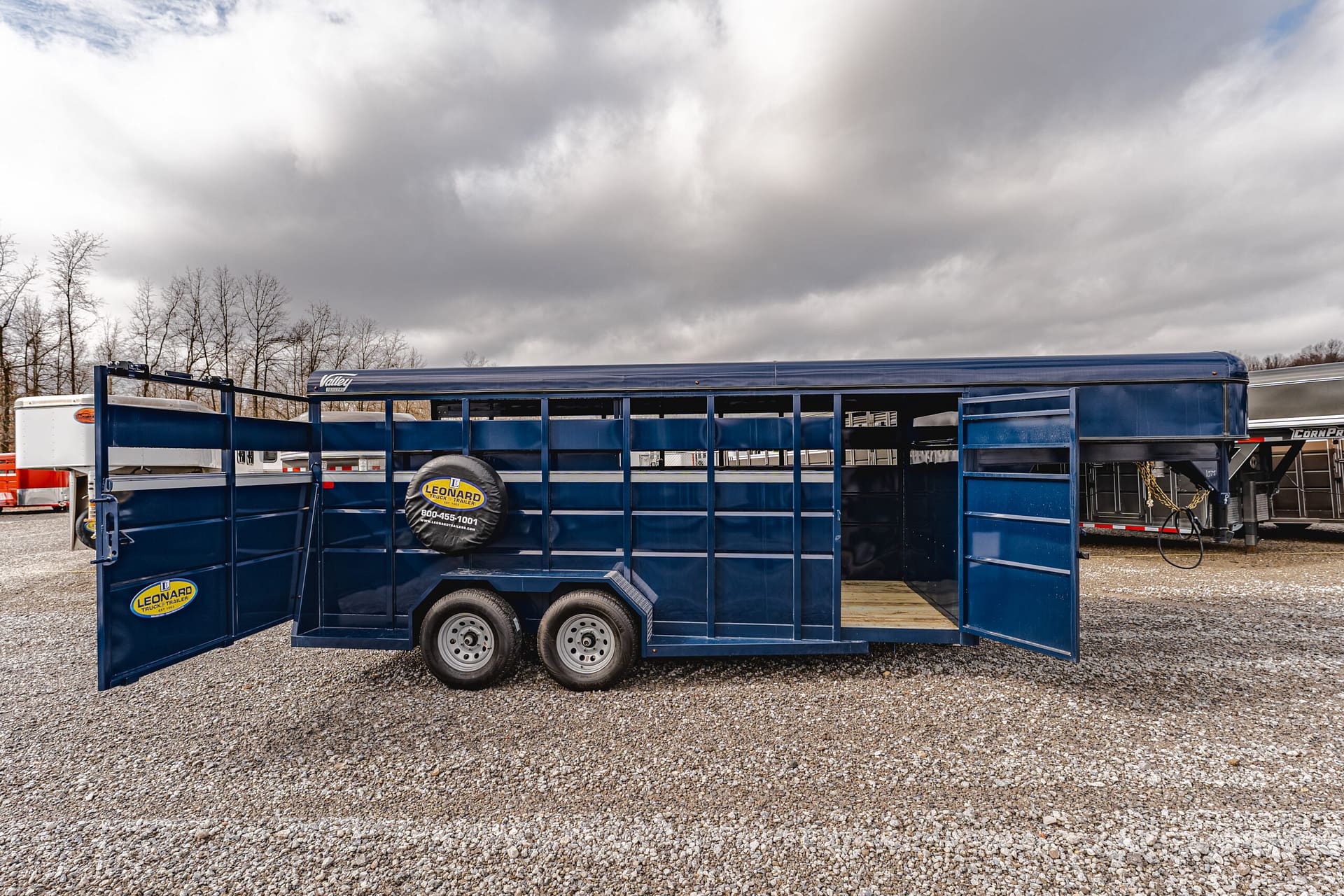 Blue livestock trailer with open rear doors on a gravel lot, cloudy sky in the background.