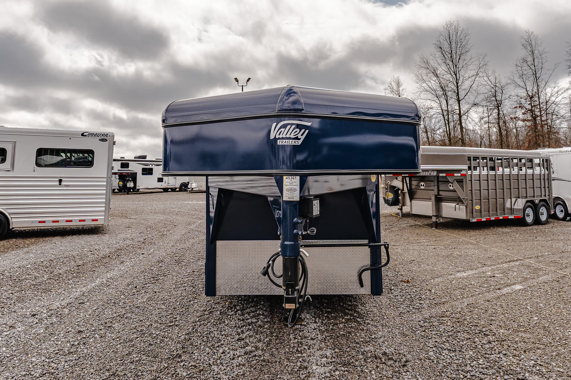 Front view of a blue Valley Trailers gooseneck trailer parked among other horse trailers on a gravel lot under a cloudy sky.