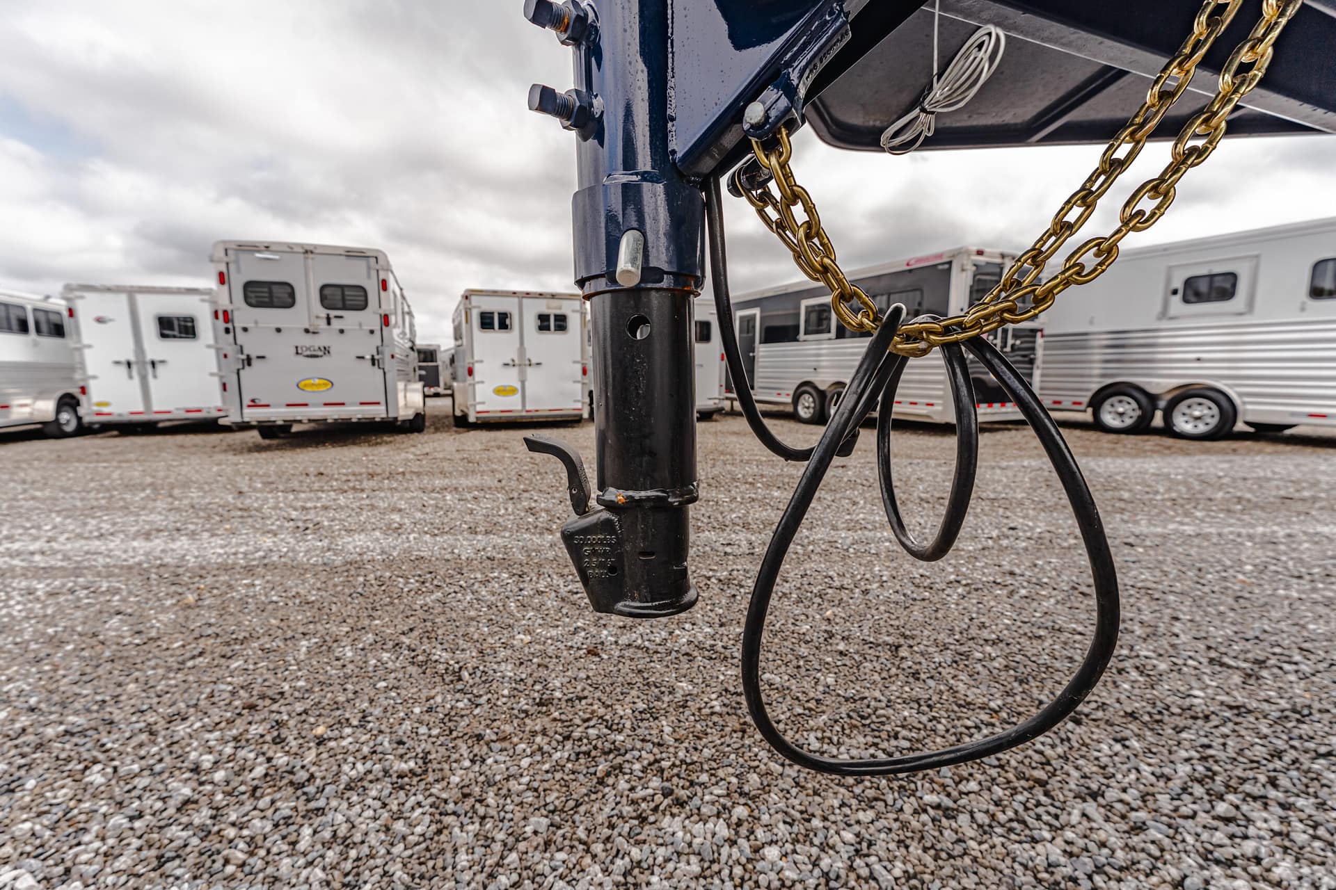 Close-up of a trailer hitch with chains and cables, with white horse trailers parked on a gravel lot in the background.