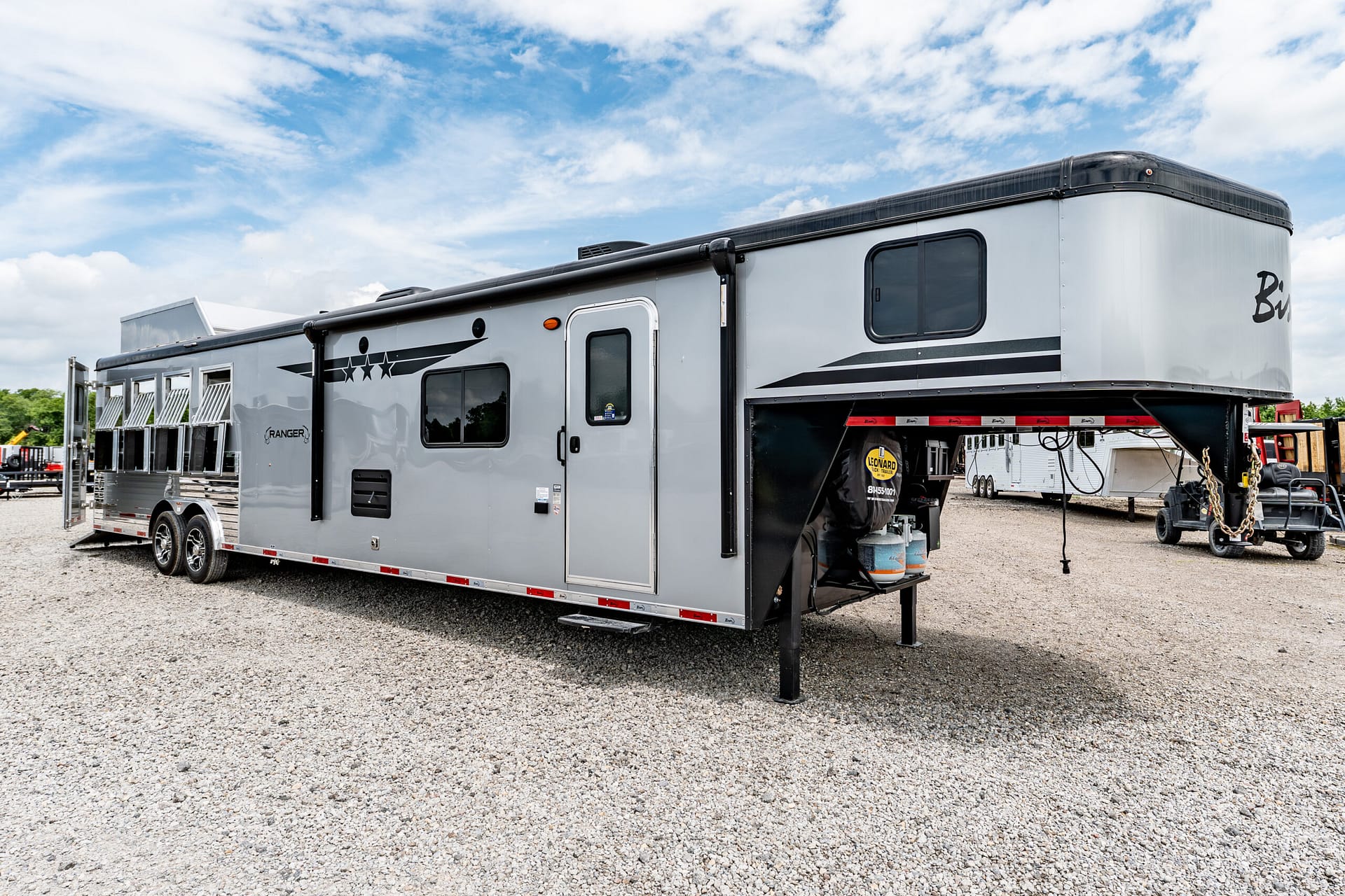 Large silver gooseneck trailer parked on a gravel lot under a partly cloudy blue sky.