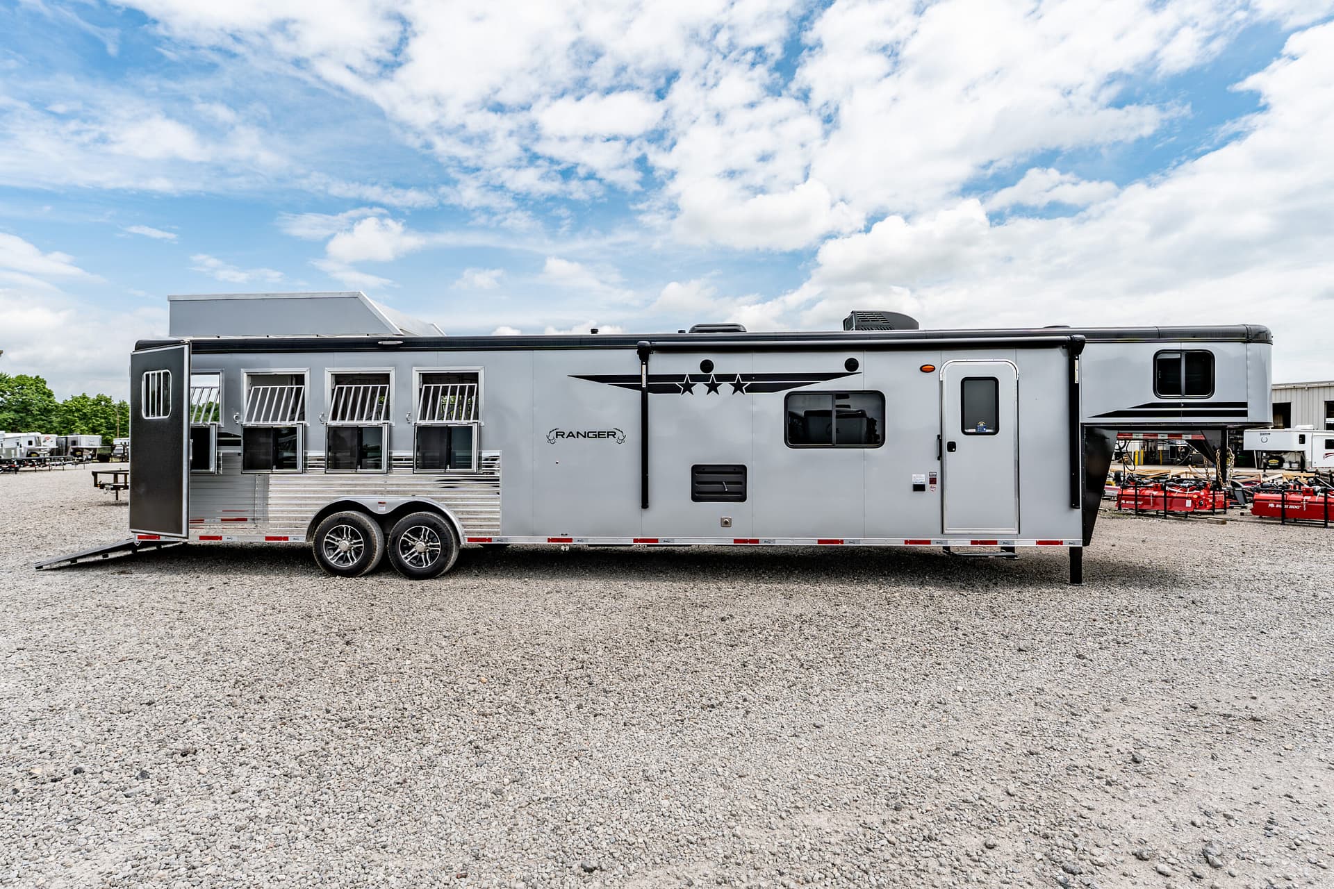 Long silver horse trailer parked on gravel with the back ramp lowered and multiple open windows along its side.