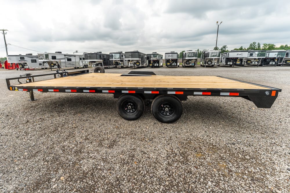 Two-axle flatbed trailer with a wooden deck sits on a gravel lot, with travel trailers lined up in the background.