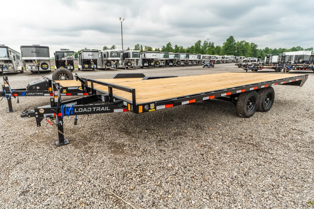 Flatbed trailer with a wooden deck and dual axles sits on a gravel lot, with a row of horse trailers in the background.