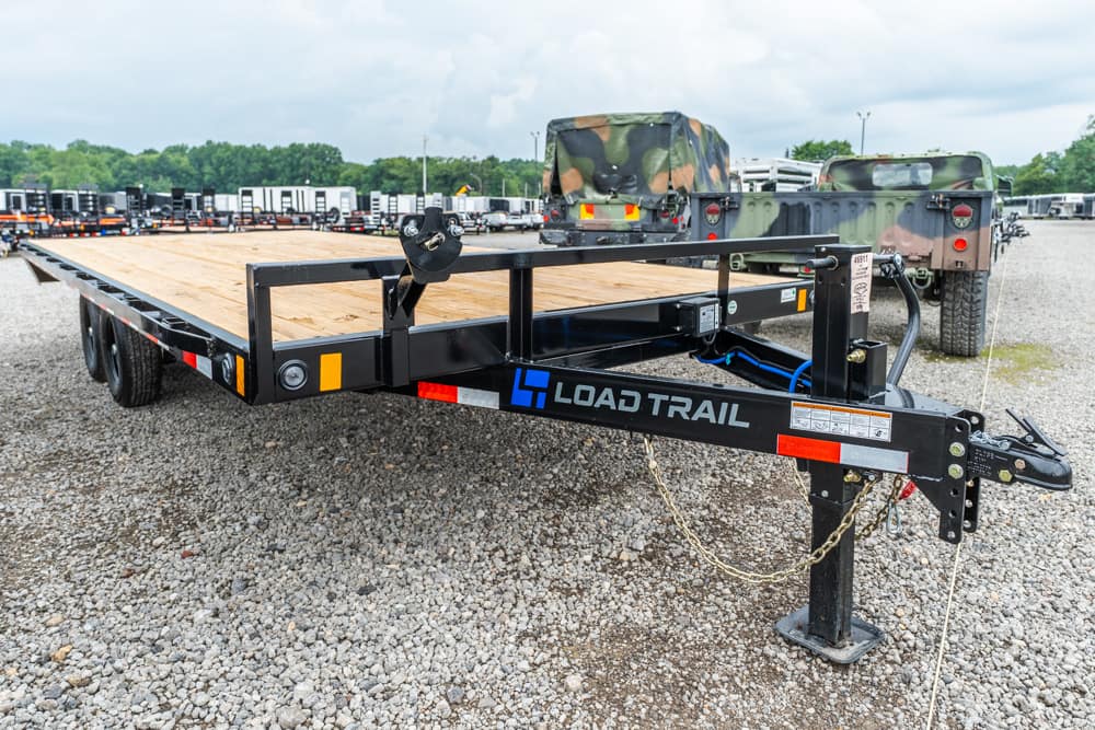 Close-up of the trailer tongue and safety chains on a black Load Trail flatbed trailer with a wooden deck, parked on a gravel lot.