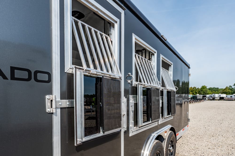 Dark aluminum trailer with multiple barred, vented windows along its side, parked on a gravel lot.