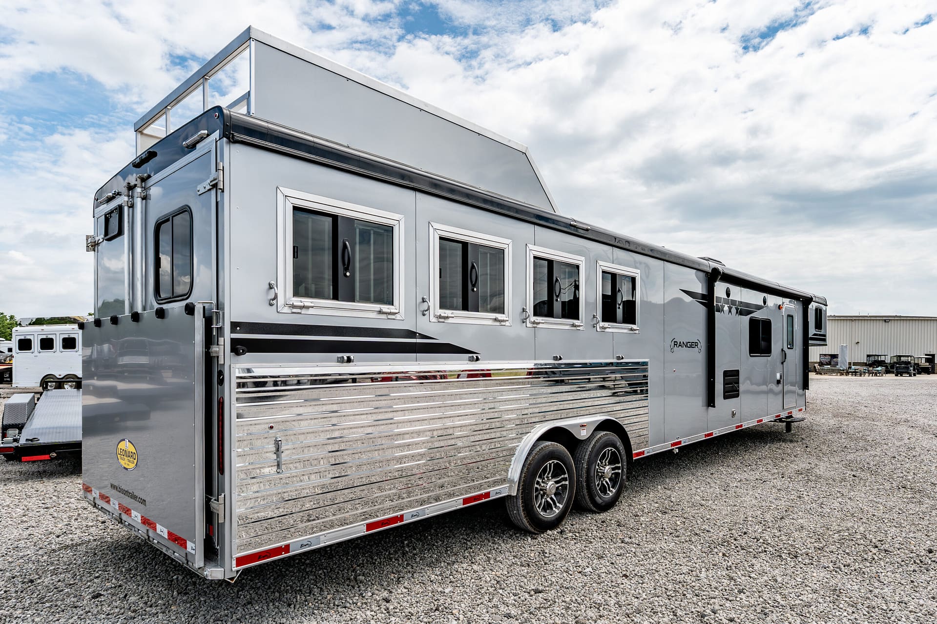 Side view of a large silver travel trailer with multiple rectangular windows and chrome trim, parked on a gravel lot under a cloudy sky.