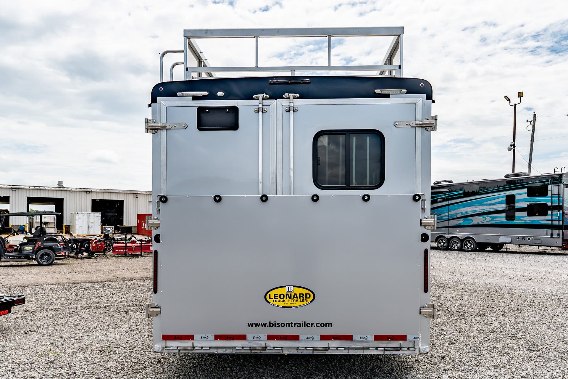 Rear view of a gray trailer with Leonard logo and www.bisontrailer.com, parked in a gravel lot with other trailers in the background.