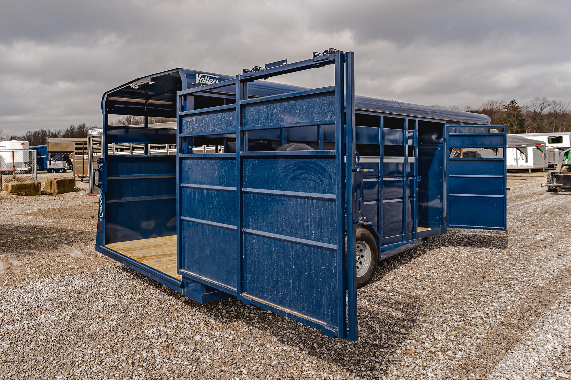 Blue metal livestock trailer with open side gate, parked on a gravel lot under a cloudy sky.