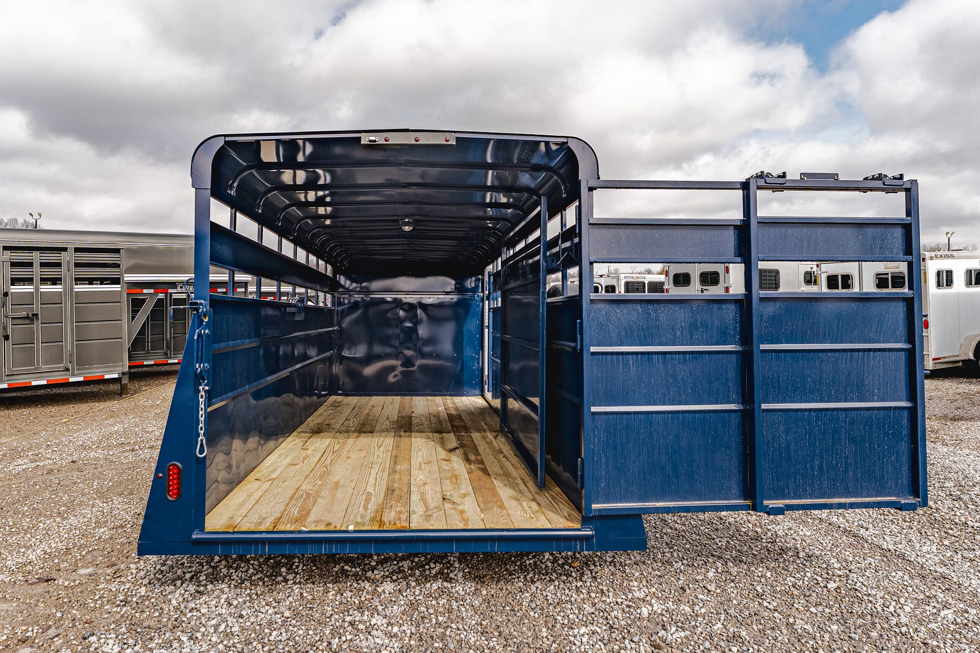 Blue livestock trailer with open rear ramp and wooden floor, parked on a gravel lot with other trailers under a cloudy sky.