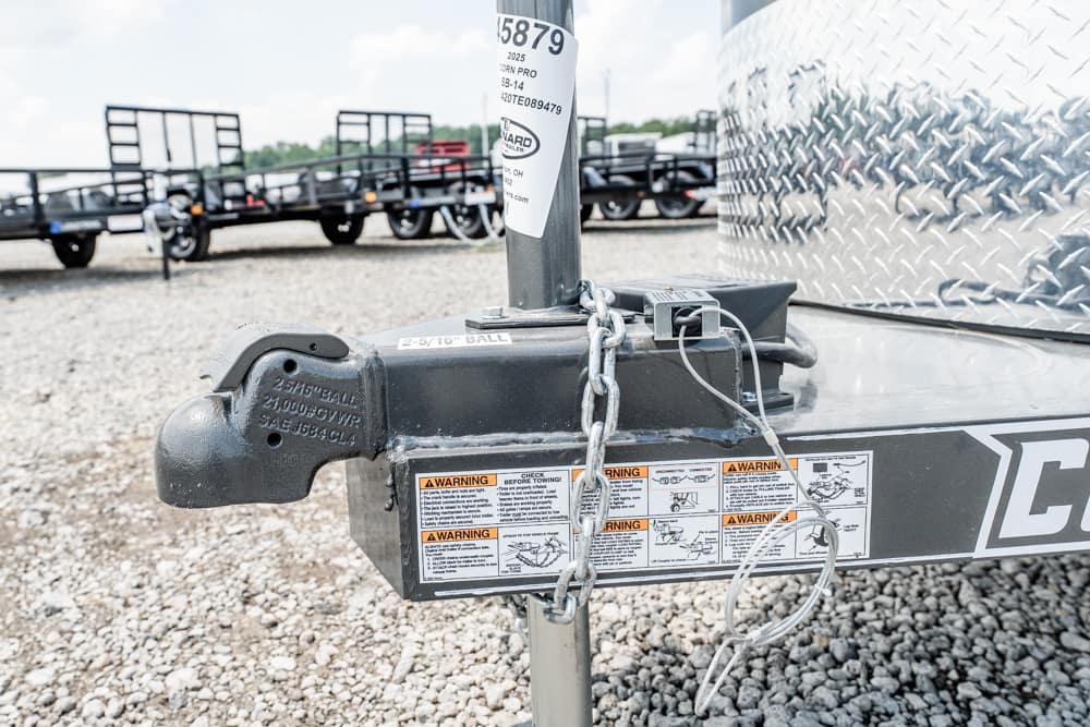 Close-up of a trailer hitch with safety chains and warning labels on a gravel lot.