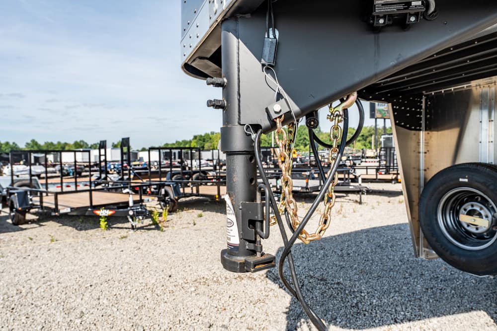 Close-up of a trailer hitch with safety chains and electrical wiring, front of a gooseneck trailer in a lot of trailers and trucks behind it.