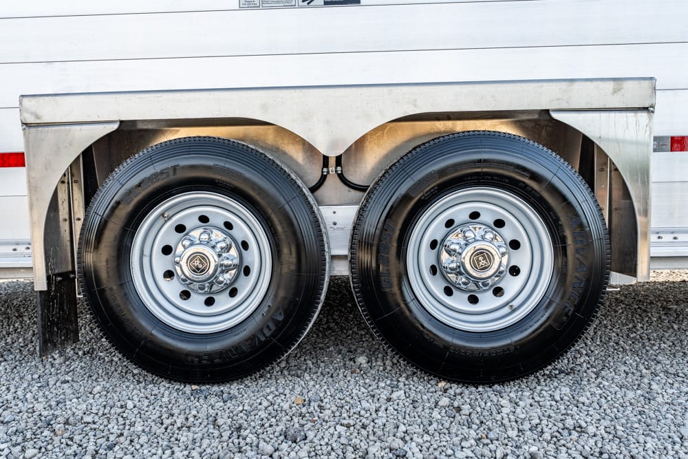 Tandem trailer wheels with black tires and shiny chrome hubcaps mounted on a metal fender over a gravel surface