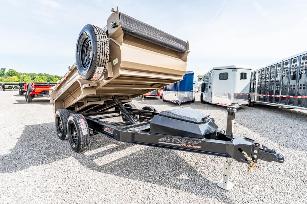 Dump trailer raised for unloading on a gravel lot, spare tire mounted on the side, with other trailers in the background.