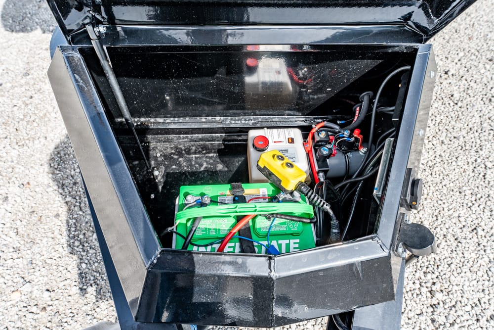 Open battery compartment showing a green battery and wiring inside a black metal box on gravel surface. A yellow control device rests atop the battery with red/black cables nearby.