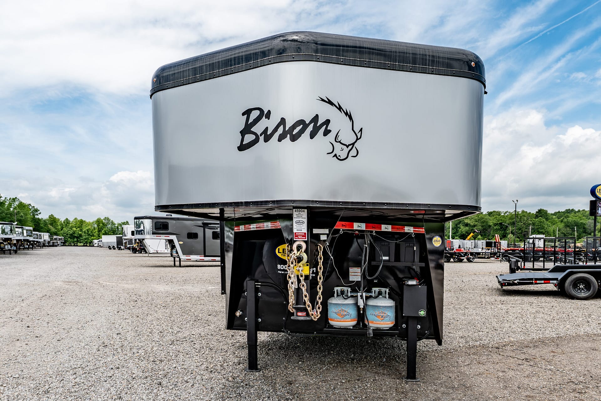 Front view of a large silver trailer in a gravel lot, with a black top edge, logo on the side, and hanging hitch chains visible at the bottom.