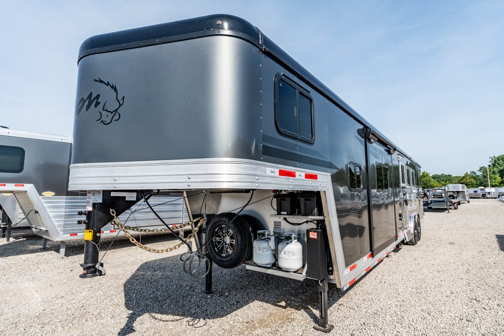 Gray gooseneck horse trailer with living-quarters, propane tanks, spare tire, and stabilizers in a gravel lot.