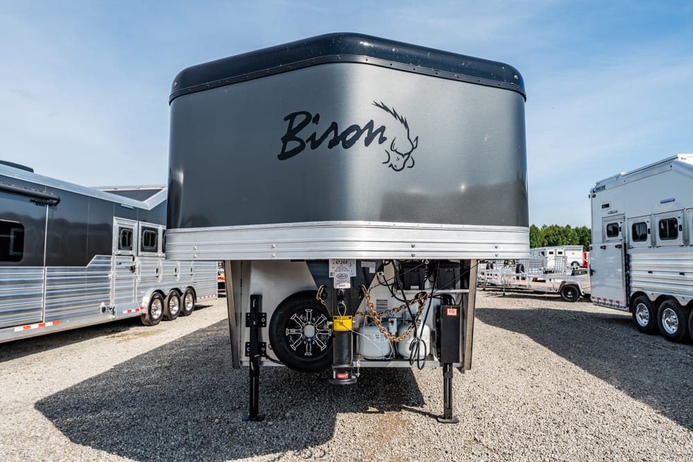 Front view of a gray Bison gooseneck horse trailer in a dealer lot, with jack stands and spare tire visible under the hitch.