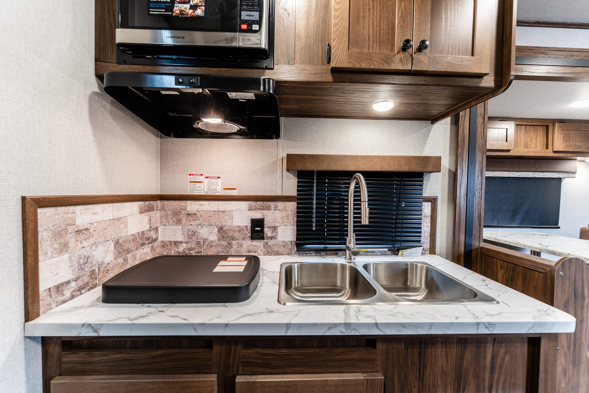 RV kitchen showing a microwave, black range hood, wooden upper cabinets, and a double stainless sink with a marble counter.