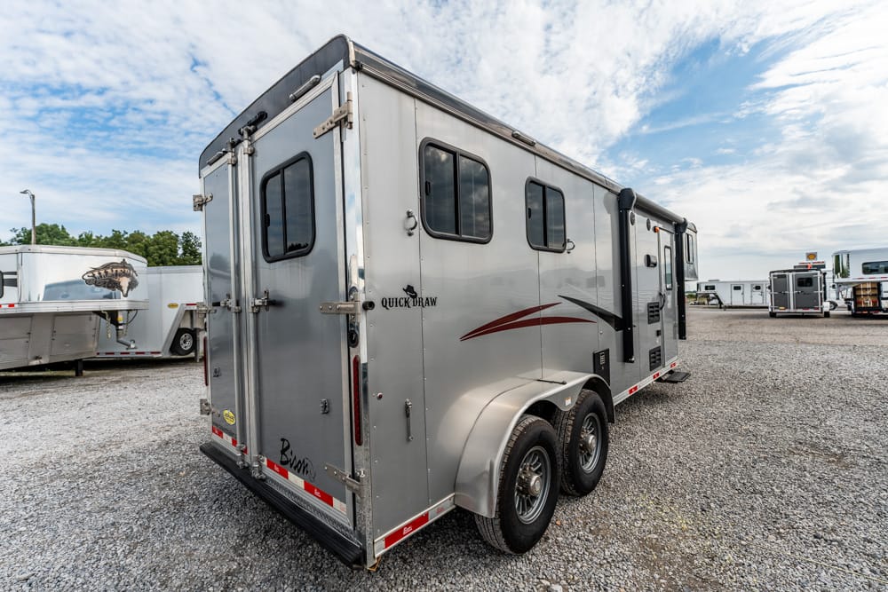 Silver double-axle horse trailer with rear doors, side windows, and decorative red/black graphics parked on a gravel lot, Quick Draw branding visible on the side.