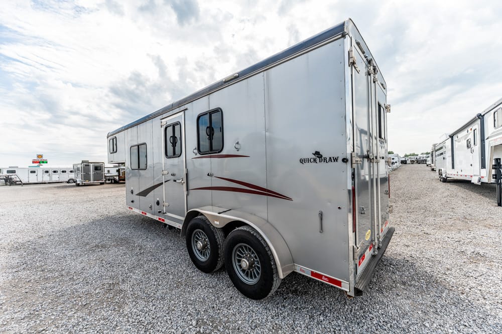 Rear three-quarter view of a silver, double-axle enclosed trailer parked on a gravel lot with other trailers in the background.