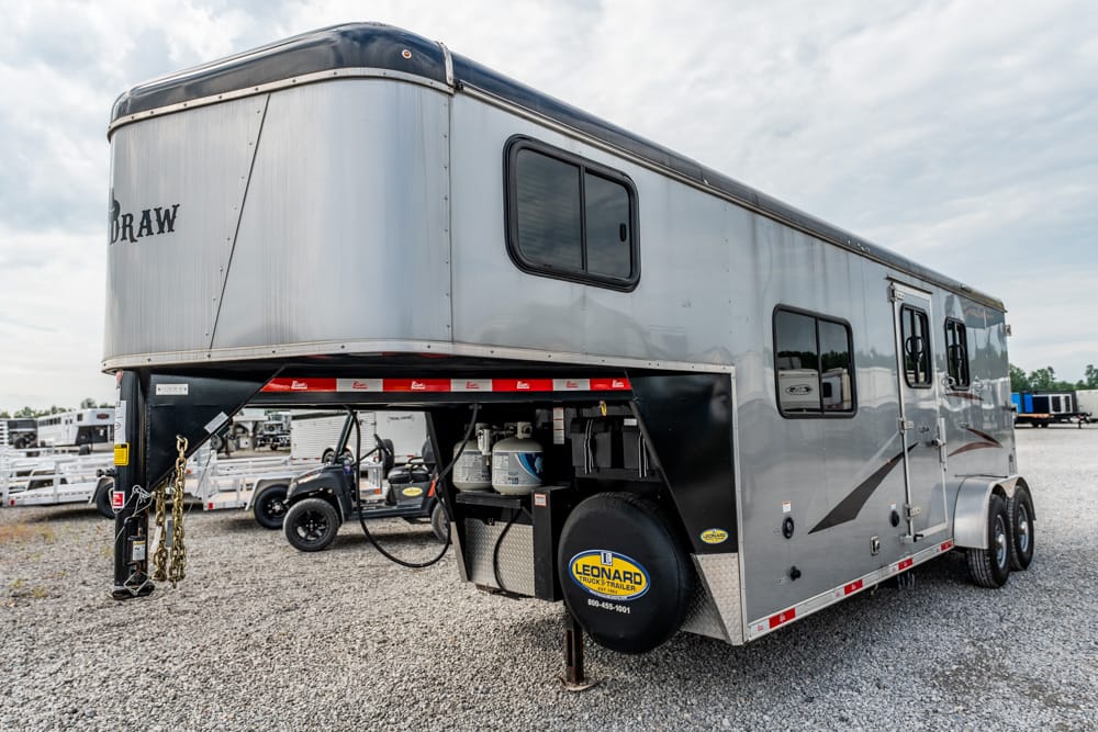 Silver fifth-wheel trailer on a gravel lot, showing the raised front hitch area, dual axles, and side windows.