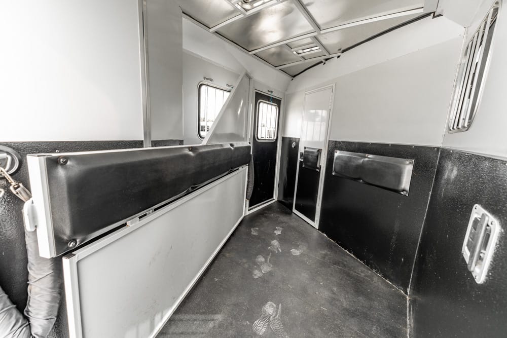 Interior of a narrow transport trailer with padded divider and black walls, showing a doorway at the far end and scuffed floor. Used as a cargo or livestock trailer interior.