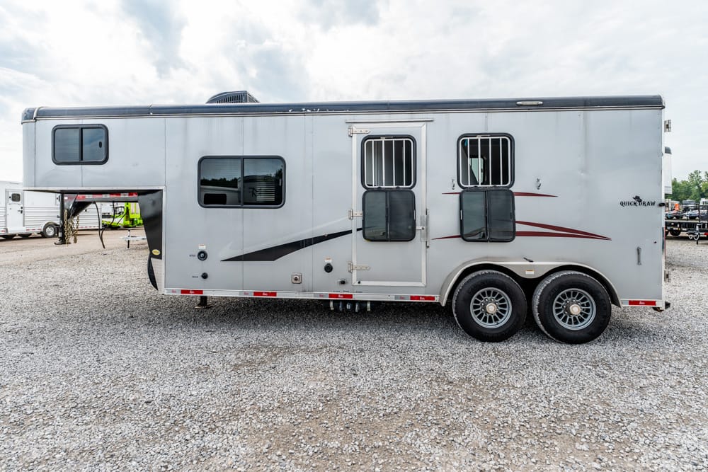 Large silver horse trailer with two axles and a gooseneck hitch parked on a gravel lot, side windows and stall bars visible.