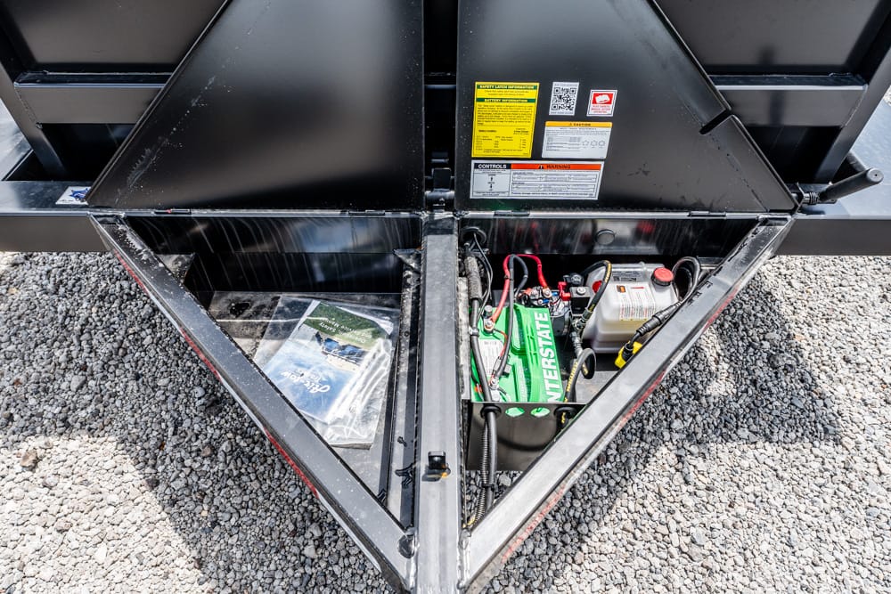 Open trailer storage with two side-by-side compartments: left shows a bag/manual, right holds a green Interstate battery and a white container on gravel surface.