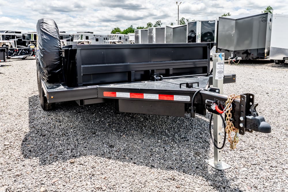 Black flatbed trailer with a spare tire mounted on the side, parked on a gravel lot among other cargo trailers.",