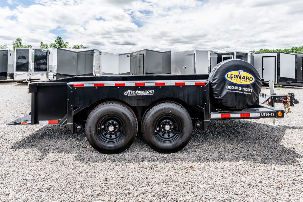 Black dual-axle utility trailer on a gravel lot, with a spare tire cover labeled Leonard and red reflective trim along the top edge of the trailer side.