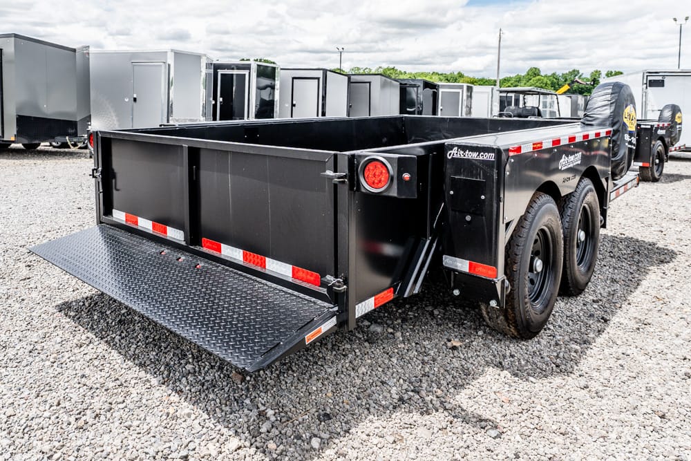 Black two-axle dump trailer with a lowered diamond-plate ramp on a gravel lot.