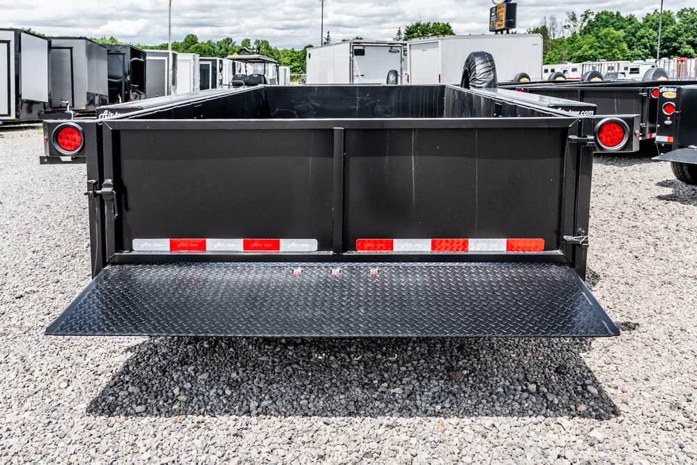 Black dump trailer viewed from the rear with the tailgate open and a diamond-plate ramp lowered to the gravel lot surface.