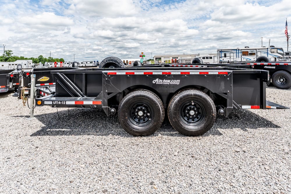 Black tandem-axle utility trailer on gravel lot, with other trailers in background; branding reads 'Hit-tow.com' on side panel.
