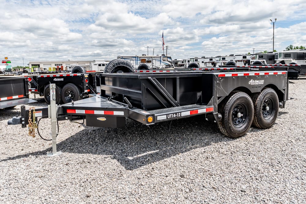 Black dual-axle dump trailer on a gravel lot with red-and-white reflective trim and spare tires, parked among other trailers in the background