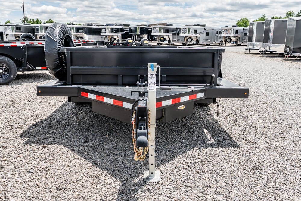 Rear view of a black utility trailer on a gravel lot, spare tire mounted to the side and safety chains hanging from the hitch, with other trailers in the background.
