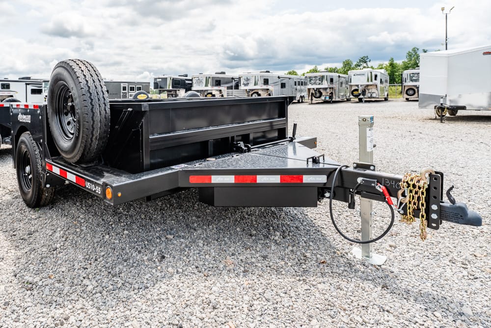 Black equipment trailer with a spare tire mounted on the left, parked on a gravel lot; front hitch area with chains and cables visible.