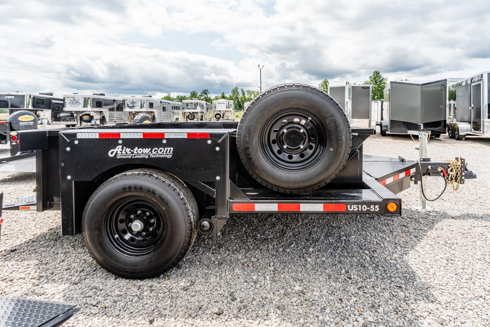 Black utility trailer with a large spare tire mounted on the side, displayed in a gravel lot with other trailers in the background.