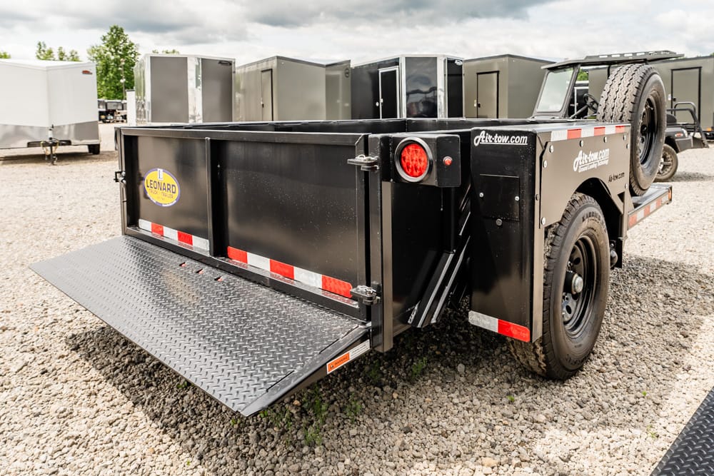 Black utility trailer with a lowered diamond-plate loading ramp, red/white safety stripes, and a spare tire mounted on the side, parked on gravel.