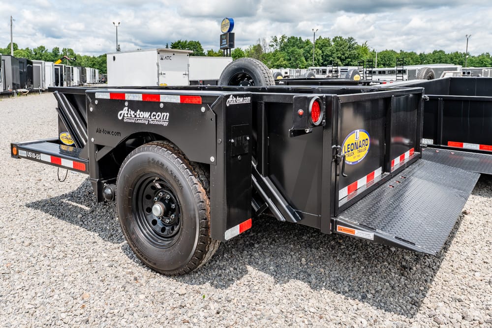 Black utility trailer with loading ramp down, spare tire on the side, and Leonard logo in a gravel lot