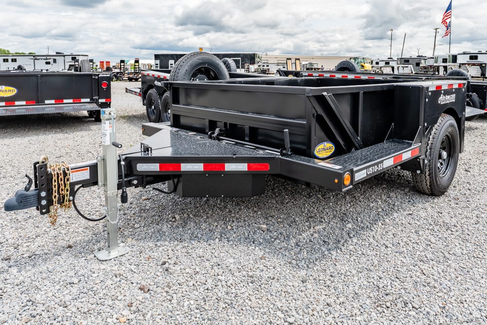 Black heavy-duty equipment trailer on a gravel lot, front hitch with chained safety chains visible and a mounted spare tire on the side.