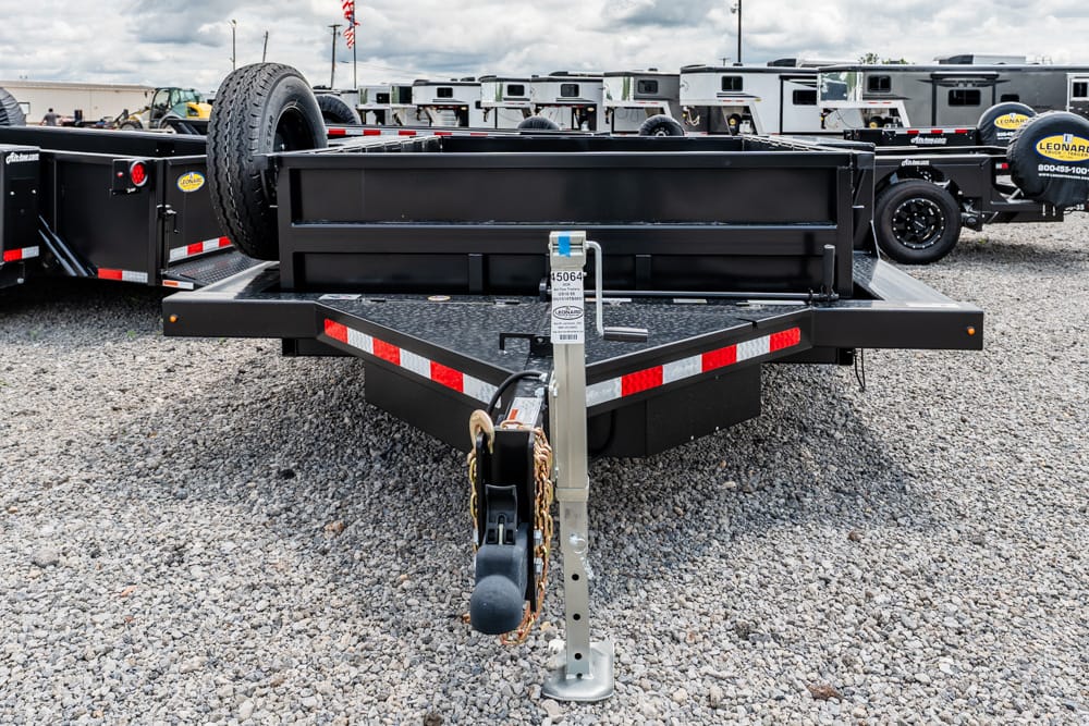 Front view of a black flatbed trailer hitch with safety chains and a spare tire, parked on a gravel lot with other trailers in the background.