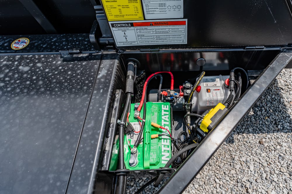 Open battery compartment with a green Interstate battery connected by red and black cables, surrounded by wiring and warning labels on a metal tray.